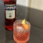 A glass of Negroni garnished with an orange peel twist beside a bottle of Campari. Campari lends the distinctive red color.