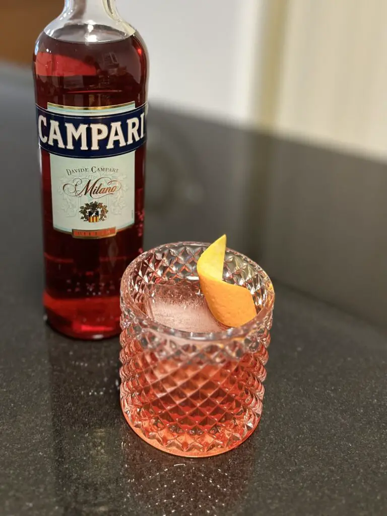 A glass of Negroni garnished with an orange peel twist beside a bottle of Campari. Campari lends the distinctive red color.